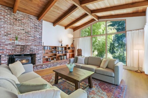 Cozy living room with a brick wall, wooden beams, large windows, and comfortable seating around a wooden coffee table.