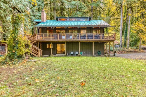 A two-story cabin surrounded by trees, featuring a green metal roof and a spacious deck with outdoor seating.