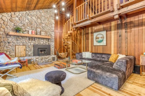 Cozy living room with stone fireplace, wooden walls, spiral staircase, and comfortable seating. Warm, inviting atmosphere.