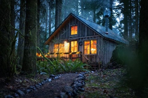 A cozy wooden cabin in a misty forest, warmly lit with glowing windows and surrounded by tall trees.