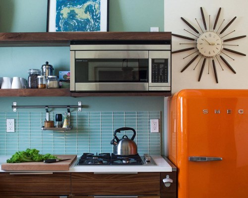 A modern kitchen featuring an orange Smeg fridge, stainless steel microwave, and a wooden countertop with fresh greens.