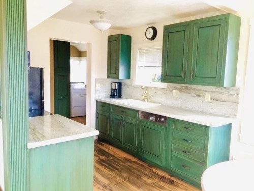 A modern kitchen featuring green cabinets, granite countertops, and wooden flooring, with a clock on the wall.
