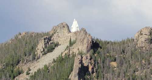 A white statue of a figure stands atop a rocky mountain, surrounded by trees and a cloudy sky.