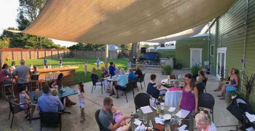 A lively outdoor gathering with people dining and children playing under a large shade sail in a backyard setting.