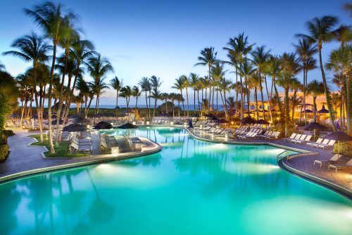 A serene pool area surrounded by palm trees, with lounge chairs and a sunset sky in the background.
