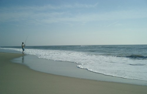 A person fishing on a sandy beach, with gentle waves lapping at the shore under a clear blue sky.