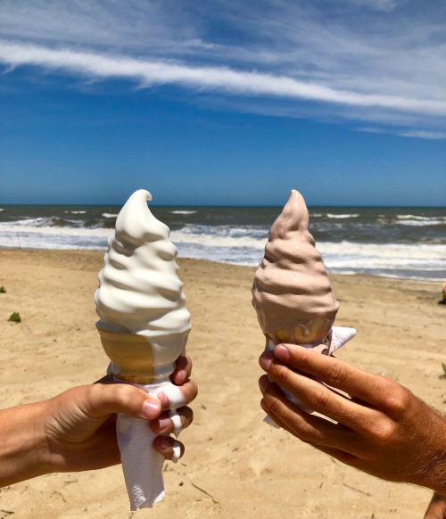 Two hands hold ice cream cones—one vanilla and one chocolate—against a beach backdrop with waves and blue sky.