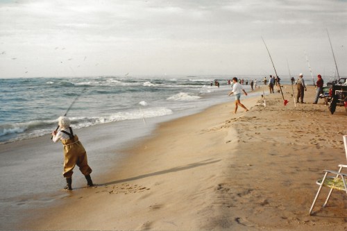A sandy beach scene with several people fishing along the shore, waves crashing and a cloudy sky overhead.