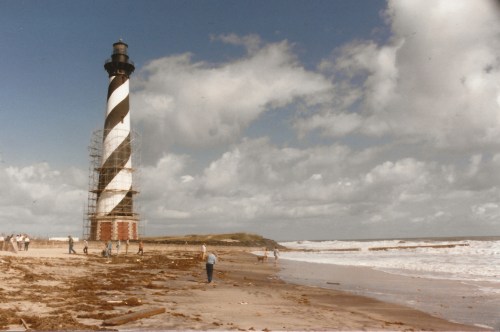 A striped lighthouse stands on a beach, surrounded by people and waves under a cloudy sky.