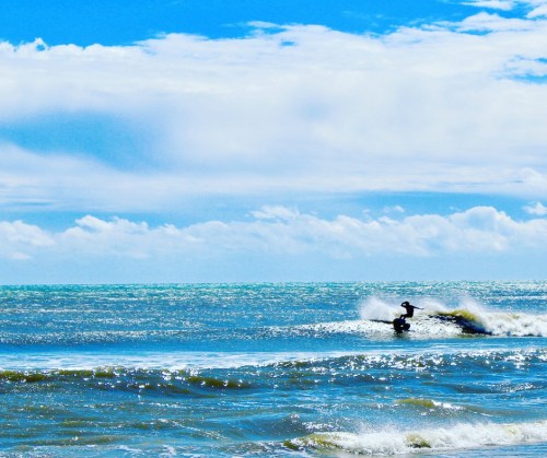 A surfer rides a wave under a bright blue sky with fluffy clouds, creating a vibrant coastal scene.