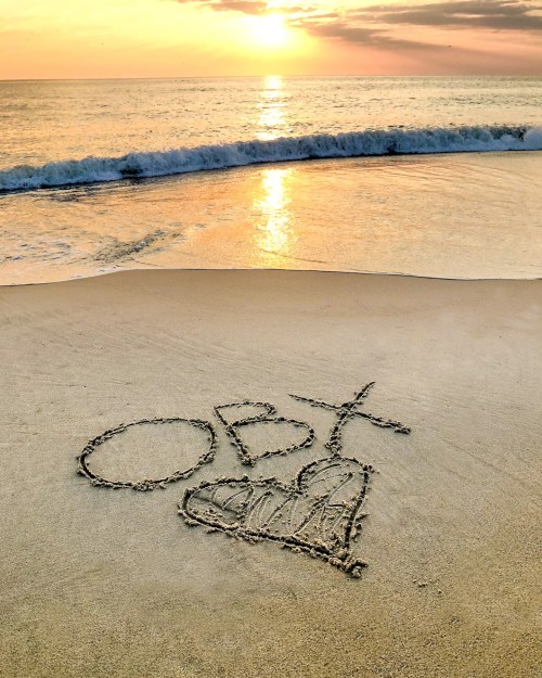 A beach at sunset with "OBY" and a heart drawn in the sand, waves gently lapping at the shore.