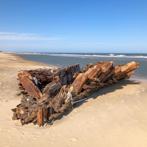 A weathered wooden shipwreck on a sandy beach, with calm waves and a clear blue sky in the background.