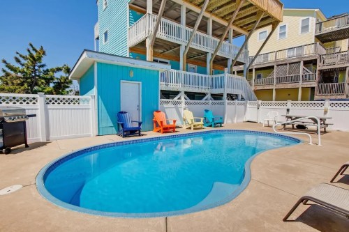 A bright pool area with colorful chairs, surrounded by a white fence and multi-story beach houses in the background.