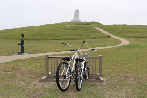 Two bicycles parked on a wooden rack, with a grassy hill and a monument in the background under a cloudy sky.