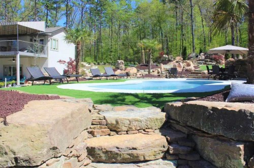 A serene backyard with a circular pool, lounge chairs, and lush greenery, surrounded by stone steps and landscaping.