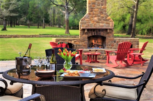 A cozy outdoor patio with a stone fireplace, wine, snacks, and red chairs surrounded by greenery.