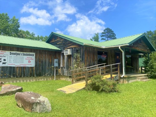 A rustic smokehouse with a green metal roof, surrounded by grass and trees, featuring a sign and a ramp for accessibility.