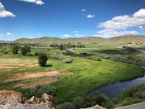 A scenic view of green fields and a winding river under a blue sky with fluffy clouds and distant hills.