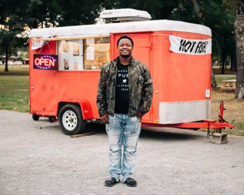A man stands in front of a red food truck with an "Open" sign, wearing a camouflage jacket and jeans.