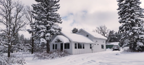 A snowy landscape featuring a white house surrounded by tall trees and a car parked nearby.