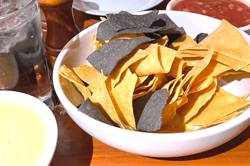 A bowl of tortilla chips, featuring yellow and blue corn varieties, alongside salsa and a drink.