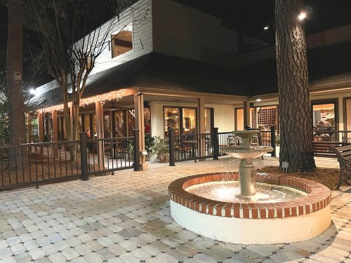 A nighttime view of a restaurant patio featuring a fountain and string lights, surrounded by trees and seating areas.