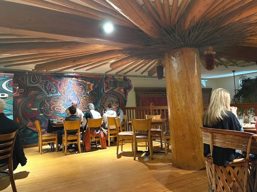 Interior of a restaurant featuring wooden beams, colorful wall art, and patrons seated at tables.