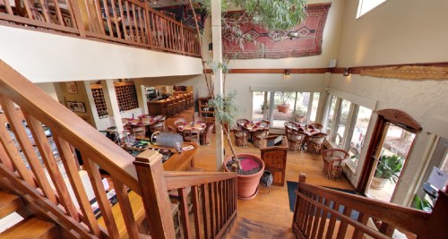 A spacious restaurant interior with wooden stairs, tables, and large windows, featuring plants and decorative artwork.