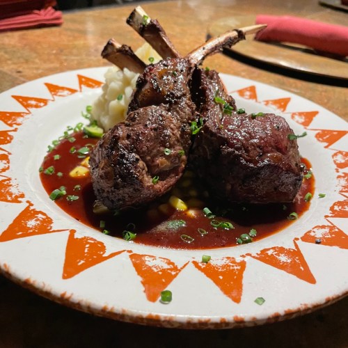 A plate of grilled lamb chops garnished with green onions, served on a bed of sauce and accompanied by mashed potatoes.