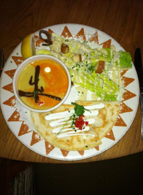 A plate with a salad, a bowl of soup, and two quesadillas, garnished with sauces and herbs.