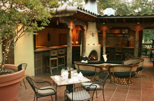 Cozy outdoor dining area with a fireplace, wooden beams, and comfortable seating surrounded by greenery.