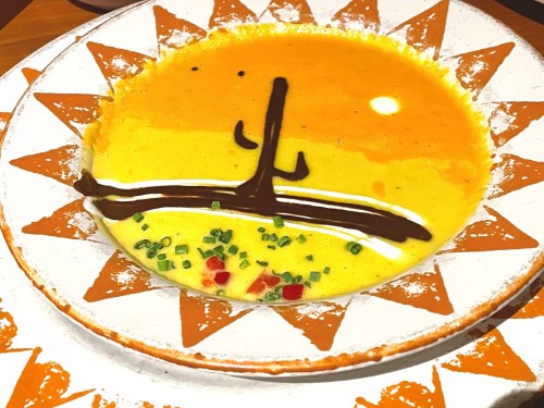 A colorful plate of soup with a sun design, featuring yellow and orange hues, garnished with green onions and red peppers.