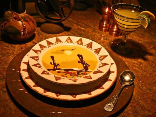 A decorative bowl of soup with artistic garnishes, accompanied by a cocktail on a rustic table setting.