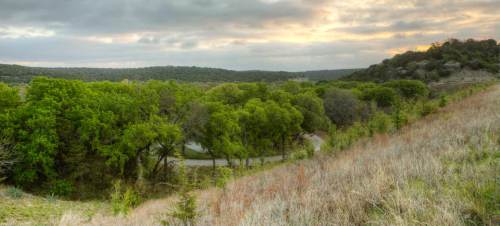 A scenic landscape featuring green trees, rolling hills, and a winding road under a cloudy sky at sunset.