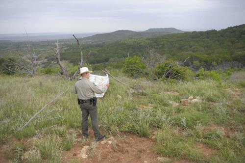 A park ranger studies a map while standing on a hillside, overlooking a lush green landscape.