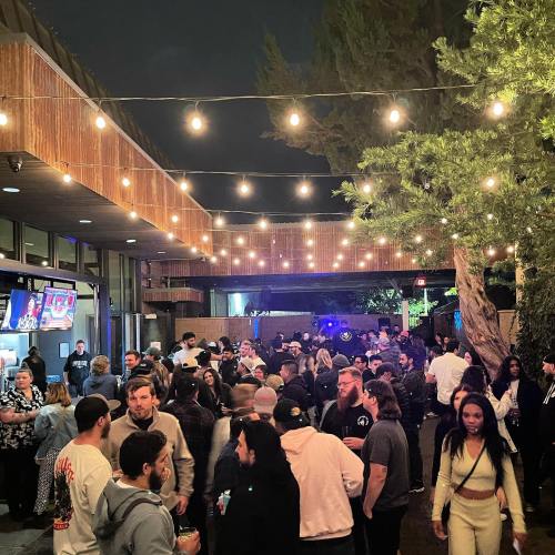 A lively outdoor gathering at night, with people socializing under string lights and a modern building in the background.