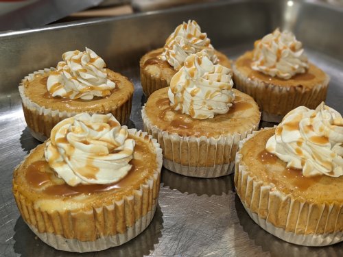 Six mini cheesecakes topped with whipped cream and caramel sauce, arranged on a metal tray.