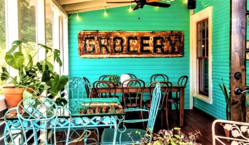 A cozy dining area with turquoise walls, vintage "Grocery" sign, and green plants, featuring metal chairs and a glass table.