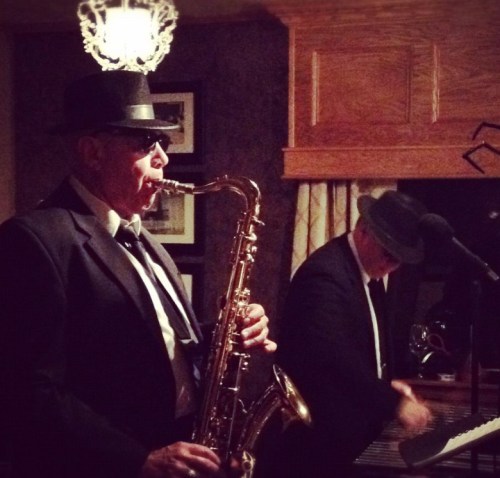 Two musicians in black suits and hats perform on stage, one playing the saxophone and the other at a keyboard.