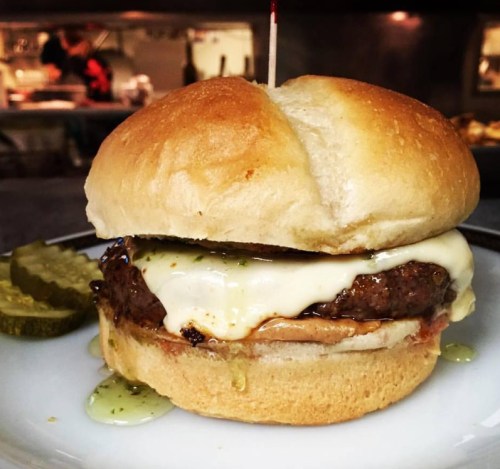 A juicy burger with melted cheese, served on a soft bun, accompanied by pickles and a drizzle of sauce.