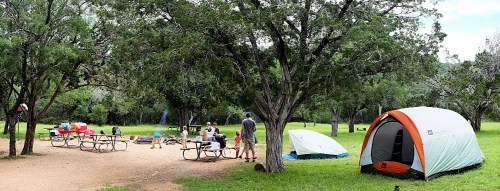 A campsite with tents and picnic tables surrounded by trees, featuring people gathered around a fire.