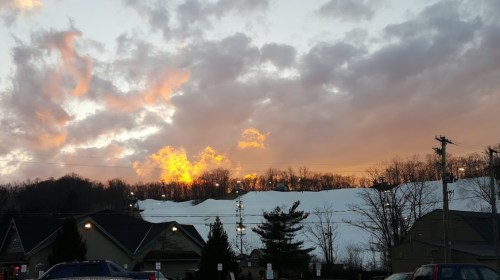 Sunset over a snowy hill, with clouds glowing orange and pink, and silhouettes of trees and buildings in the foreground.