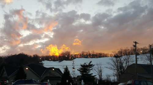 Sunset over a snowy hill, with clouds glowing orange and pink, and silhouettes of trees and buildings in the foreground.