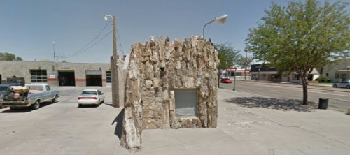 A small, rock-covered building stands on a concrete lot, surrounded by trees and parked cars under a clear blue sky.