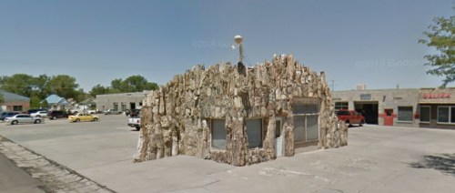 A unique building with a rocky exterior stands in a parking lot, surrounded by other structures and vehicles.