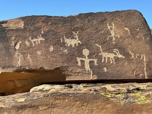 Petroglyphs on a rock surface depicting human figures and animals against a clear blue sky.