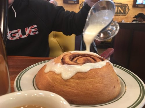 A person pours icing over a large cinnamon roll on a plate, with a cup of coffee in the foreground.