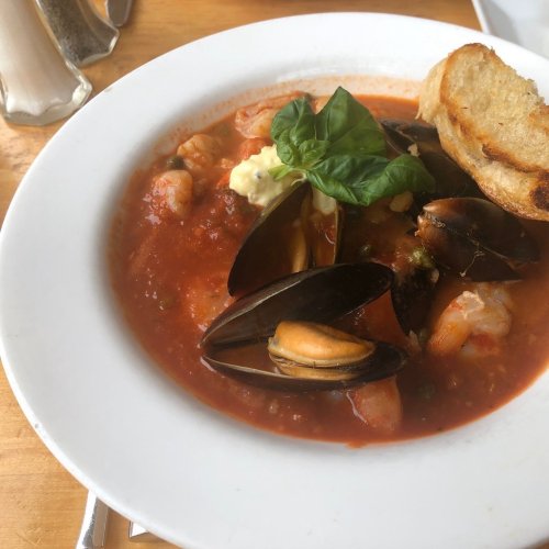 A bowl of seafood stew with shrimp, mussels, and herbs, served with a slice of toasted bread on the side.