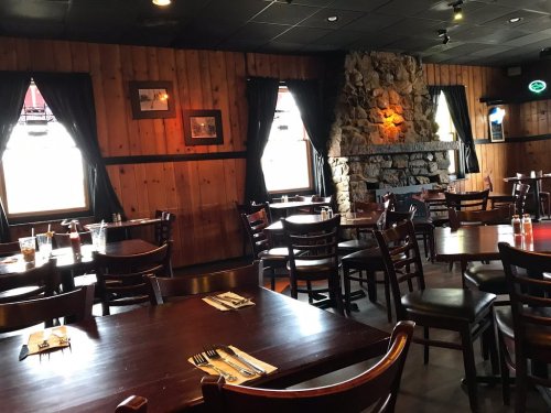 Cozy restaurant interior with wooden walls, empty tables, and a stone fireplace, featuring large windows and dark curtains.