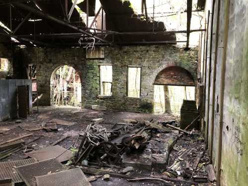 An abandoned building interior with crumbling walls, scattered debris, and overgrown vegetation visible through broken windows.